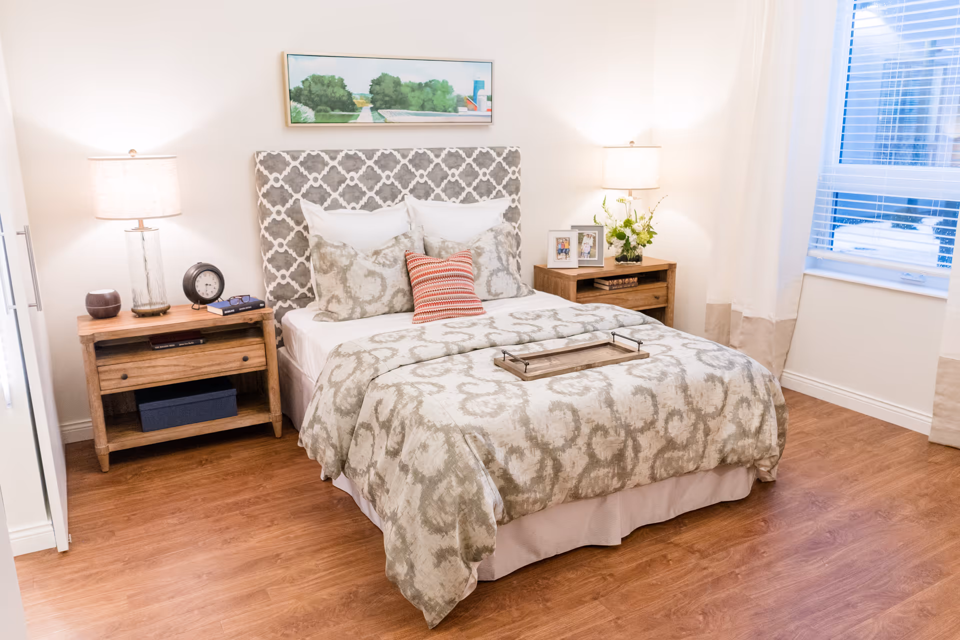 A neatly made bed with patterned bedding and multiple pillows in a bright bedroom. There are two wooden nightstands on either side of the bed, each with a lamp. One nightstand has a clock and books, while the other has framed photos and a vase with flowers. A window with white curtains is on the right side, and a landscape painting hangs above the bed.