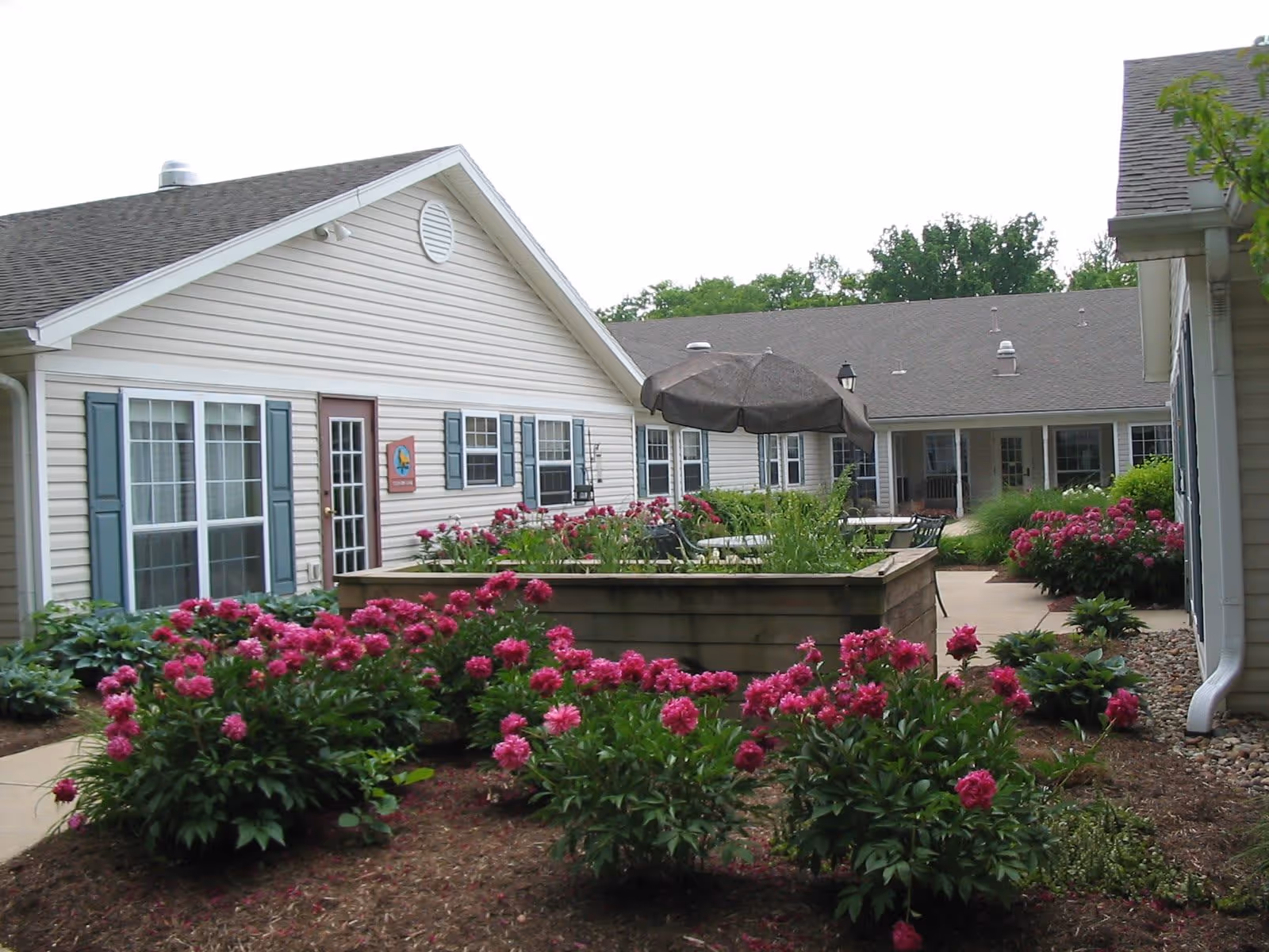 Outdoor courtyard area of a senior living facility with blooming pink flowers, a raised garden bed, patio tables with umbrellas, and beige buildings with blue shutters surrounding the space.