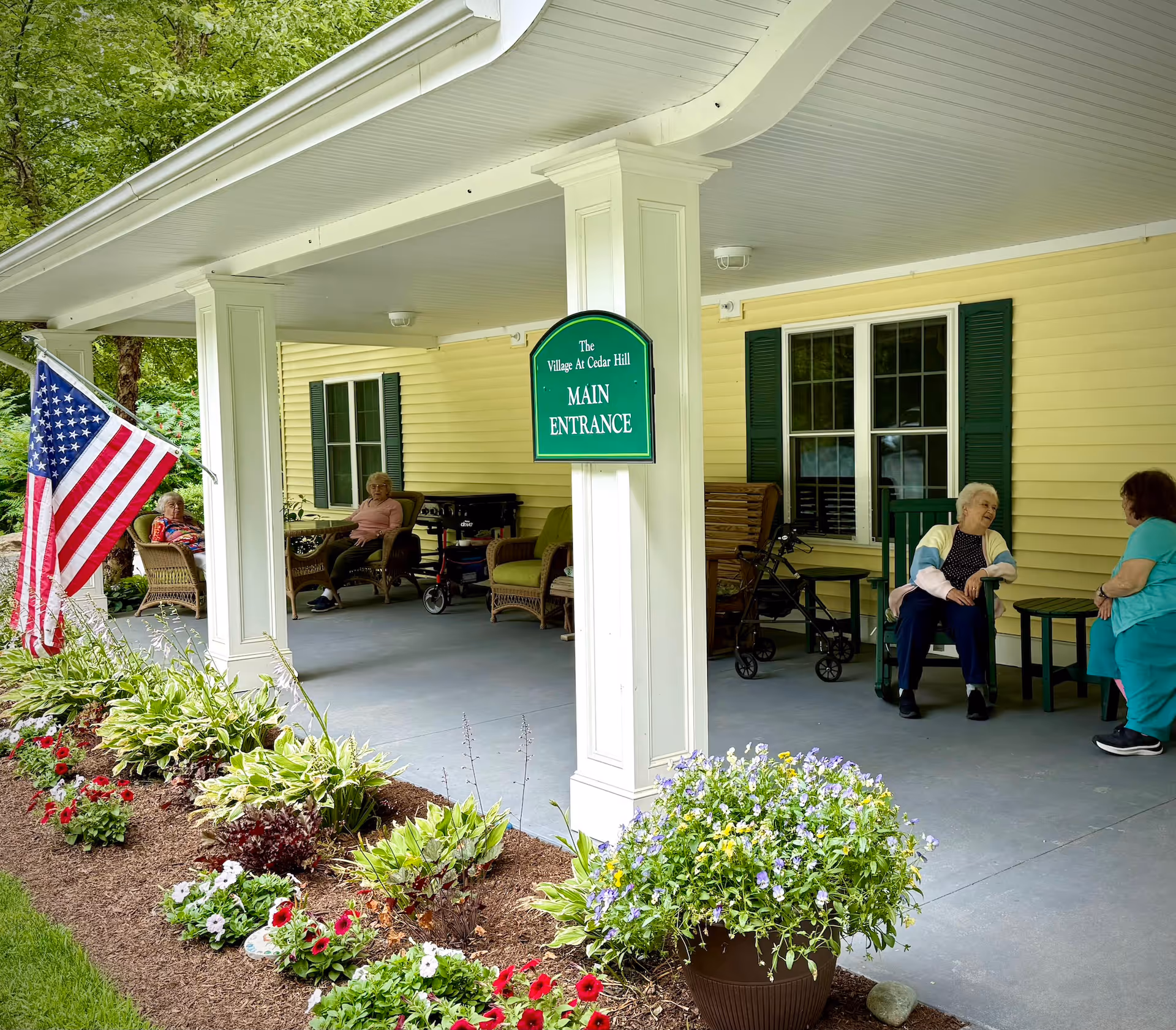 Porch area of The Village At Cedar Hill with a green sign reading 'Main Entrance' on a white pillar. Several elderly people are seated on chairs and rocking chairs under the covered porch. There is an American flag on the left side and a flower bed with colorful flowers in front of the porch. The building has yellow siding and green shutters on the windows.