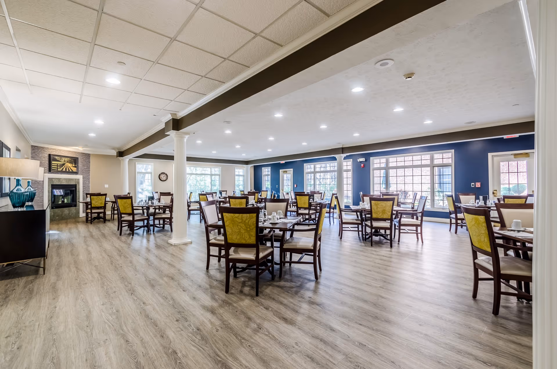 A spacious and well-lit dining room with multiple wooden tables and chairs arranged neatly. The room features large windows allowing natural light to fill the space, a fireplace on the left side, and light-colored wood flooring. The walls are painted in neutral tones with a dark blue accent wall in the background.