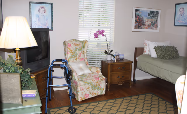 A cozy assisted living bedroom featuring a floral armchair, walker, TV, nightstand with an orchid, and a single bed by a window with blinds.