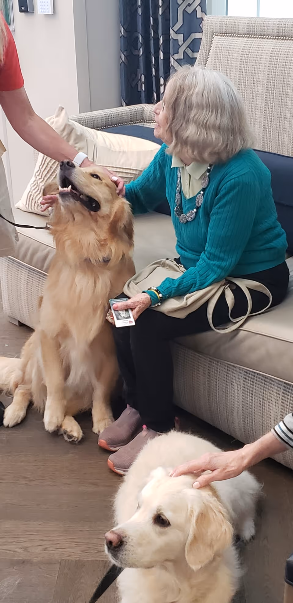 An elderly woman sitting on a cushioned bench indoors, petting a golden retriever while another person pets a second golden retriever sitting on the floor nearby.