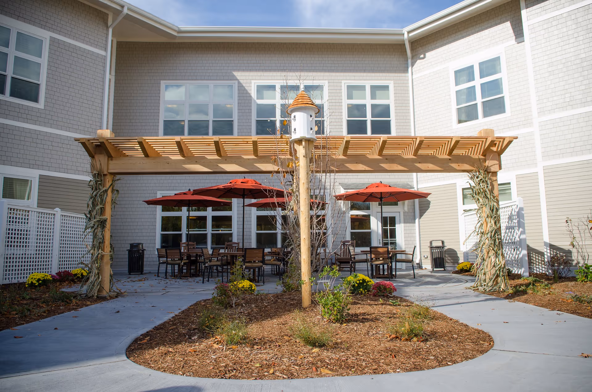 Outdoor patio area at Bridges by EPOCH at Trumbull featuring a wooden pergola decorated with dried corn stalks, several tables with red umbrellas, chairs, and a birdhouse mounted on a wooden post in the center. The patio is surrounded by a building with multiple windows and a clear sky above.