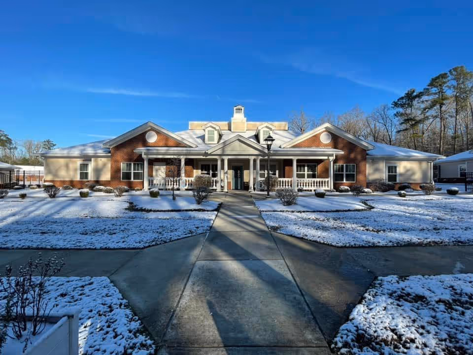 Front exterior view of a single-story brick and beige building with a covered porch and white railings, surrounded by snow-covered ground and leafless bushes under a clear blue sky.