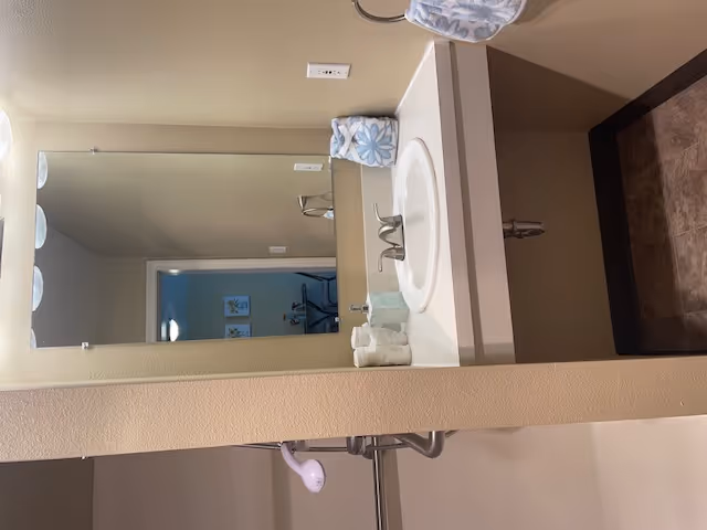 A bathroom sink area with a large mirror above the countertop. The countertop has a white sink with a silver faucet, a soap dispenser, and a folded hand towel with a blue floral pattern. The wall and countertop are beige, and the floor has brown tiles. A shower curtain rod and part of a showerhead are visible to the left.