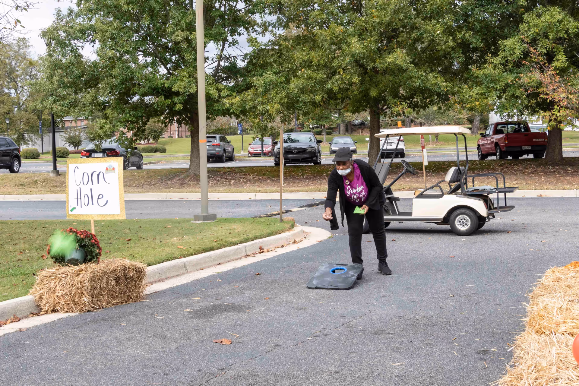 An elderly woman wearing a mask and a hat is playing cornhole outdoors on a paved area. She is tossing a bean bag towards a cornhole board placed on the ground. There is a sign that reads 'Corn Hole' next to a hay bale with a potted plant on it. In the background, there are several parked cars, trees, and a golf cart.