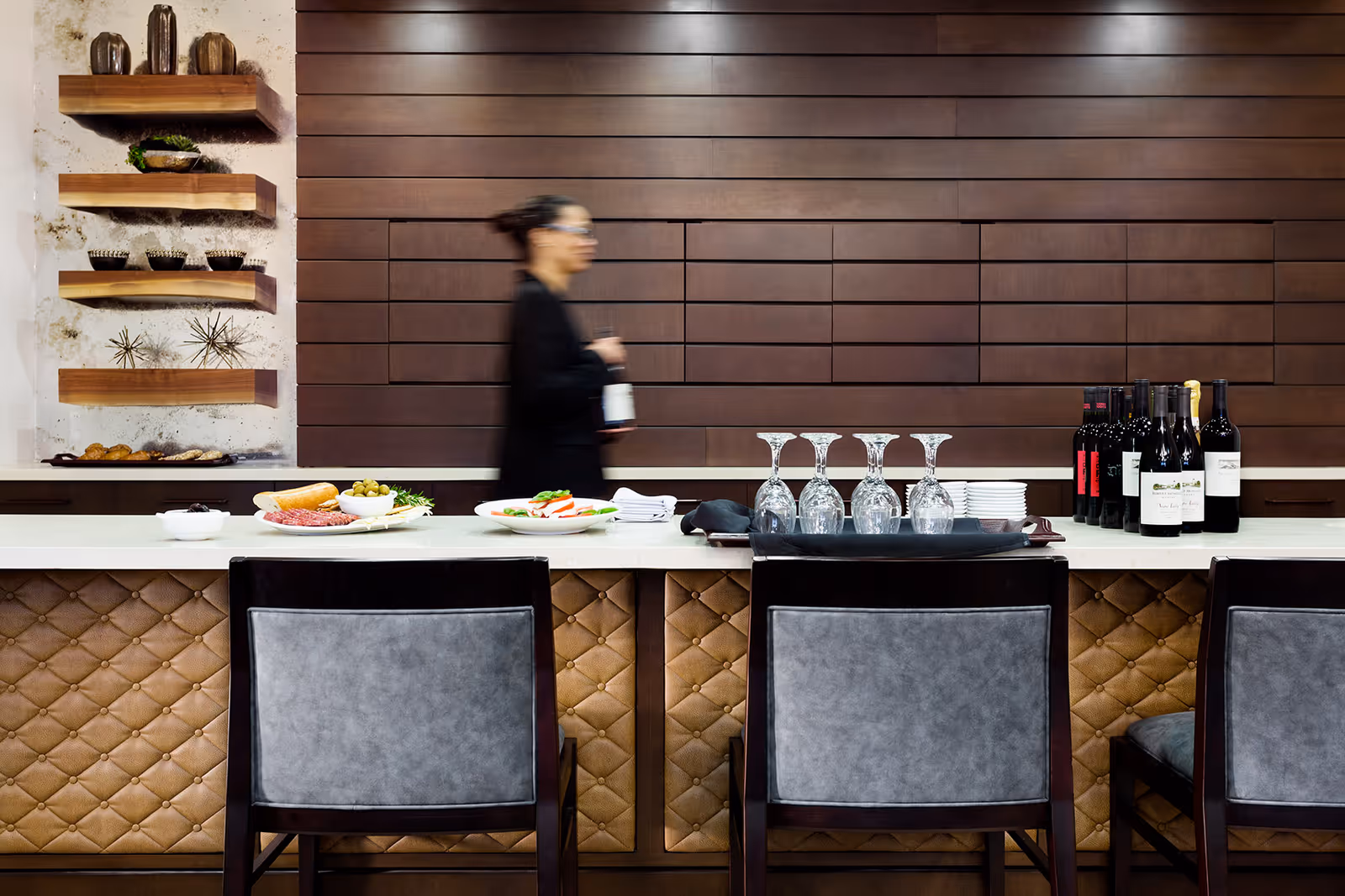 Bar-style dining counter with three chairs, wine bottles and glasses on the countertop and a blurred staff member walking behind it.