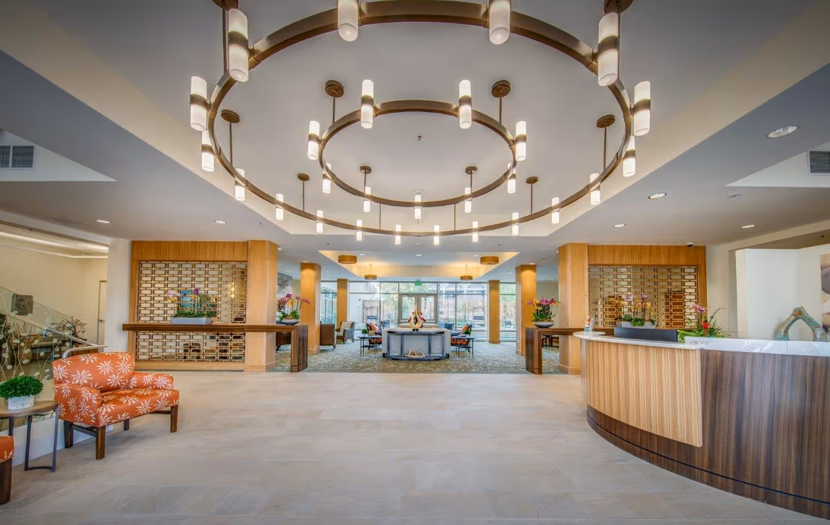 Spacious and modern senior living facility lobby with a large circular chandelier hanging from the ceiling, a curved wooden reception desk on the right, orange patterned armchairs on the left, decorative plants, and seating areas in the background near large windows letting in natural light.