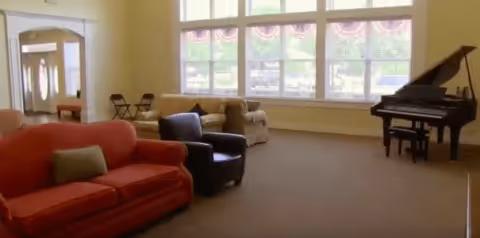 A spacious living room area with a red sofa, a black armchair, and a beige armchair arranged near large windows with decorative valances. A grand piano is positioned on the right side of the room. The room has light-colored walls and carpeted flooring.
