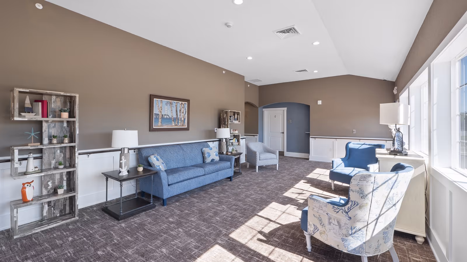 Sunlit sitting room with blue sofas and armchairs, shelving, lamps, and large windows.