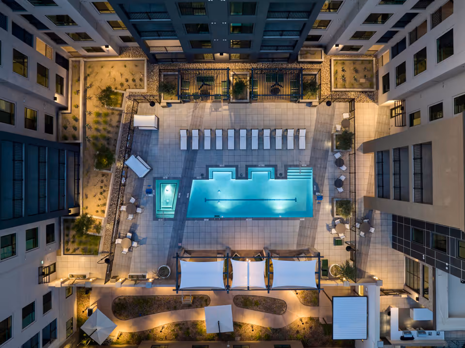 Aerial view of an outdoor courtyard at night featuring a rectangular swimming pool with adjacent hot tub, surrounded by lounge chairs, tables with umbrellas, and landscaped garden areas within a multi-story building complex.