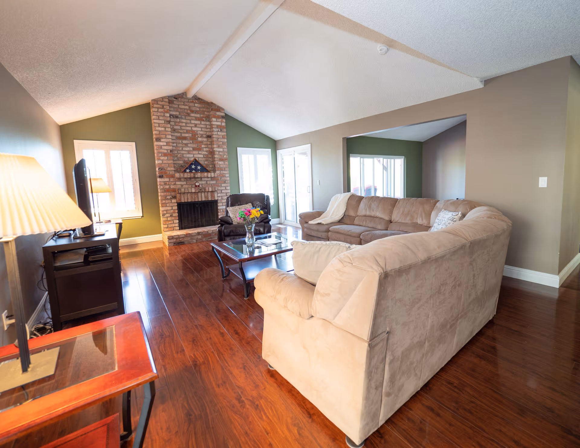 Spacious living room with a large beige sectional sofa, brick fireplace, coffee table, TV and hardwood floors.