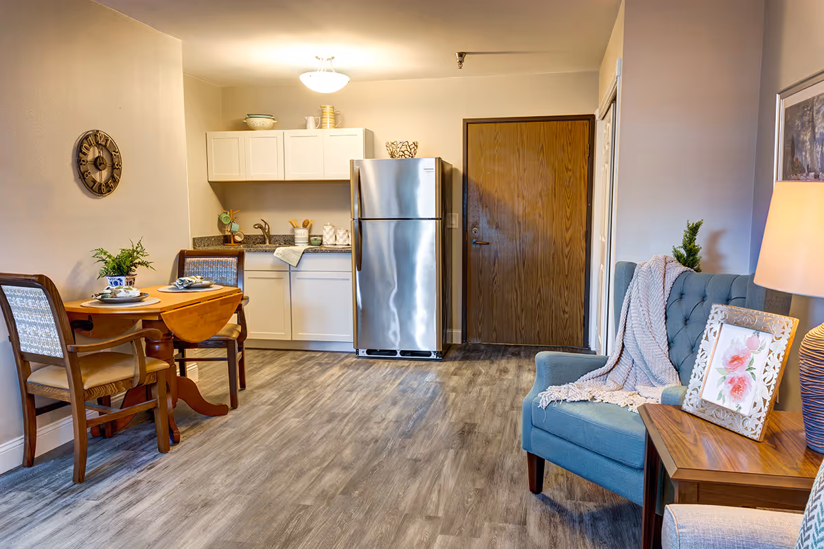 Cozy studio interior with a small kitchenette and stainless steel refrigerator, a set dining table, and a blue upholstered chair beside a side table.