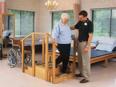 An elderly woman using a small wooden step exercise apparatus with handrails in a room, assisted by a male caregiver. The room has large windows with blinds, a wheelchair, and beds with pillows in the background.