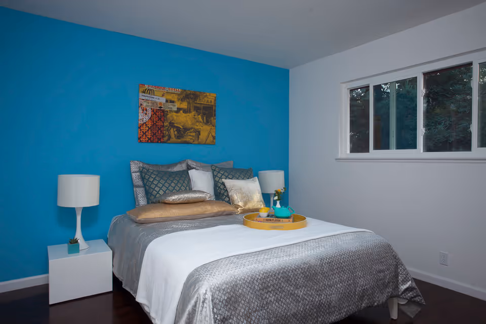 A neatly made bed with multiple pillows and a silver-gray bedspread in a bedroom with a bright blue accent wall. There is a white nightstand with a white lamp and a small plant on the left side of the bed. A colorful framed artwork hangs on the blue wall above the bed. A window with multiple panes is on the right wall, showing greenery outside.