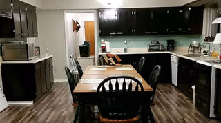 A kitchen and dining area with dark wood cabinets, a wooden dining table with six black chairs, and various kitchen appliances including a microwave, toaster, and stove. The floor has wood-patterned vinyl or laminate flooring, and there is a doorway leading to another room.