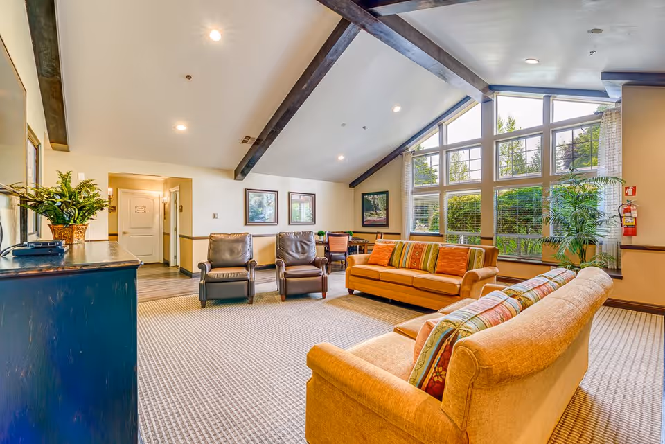 A bright and spacious living room with high vaulted ceilings and exposed wooden beams. The room features two beige sofas with colorful cushions, two brown leather armchairs, a large window with a view of greenery outside, framed artwork on the walls, a potted plant near the window, and a blue cabinet with a plant on top.
