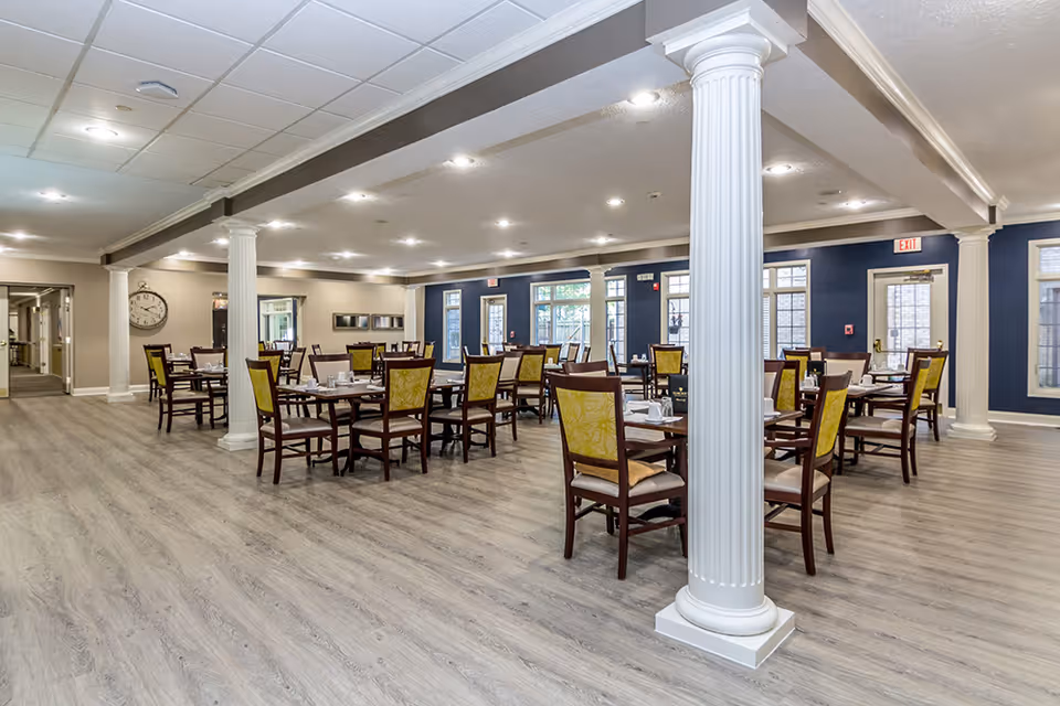 Spacious dining room in West Lafayette Assisted Living with multiple wooden tables and chairs featuring yellow cushioned backs, light wood flooring, white decorative columns, and large windows allowing natural light.