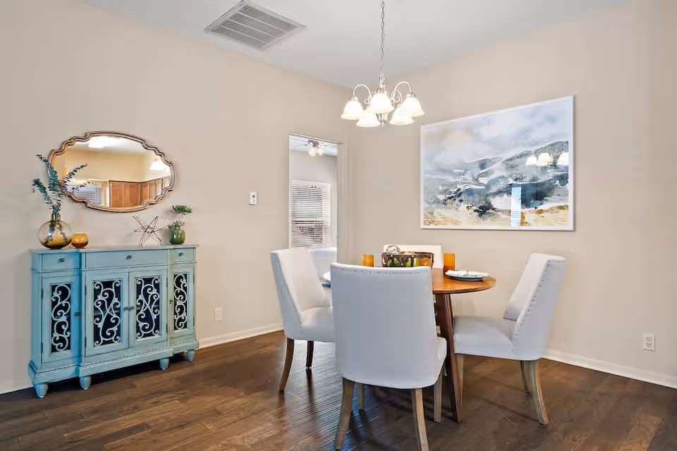 A dining room with a round wooden table surrounded by four upholstered chairs. Above the table hangs a chandelier with five lights. On the wall behind the table is a large abstract painting. To the left, there is a blue sideboard with decorative items and a mirror above it. The room has wooden flooring and beige walls.