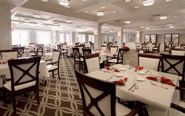 Bright, elegant dining room with multiple tables set with white linens, red napkins, and glassware.