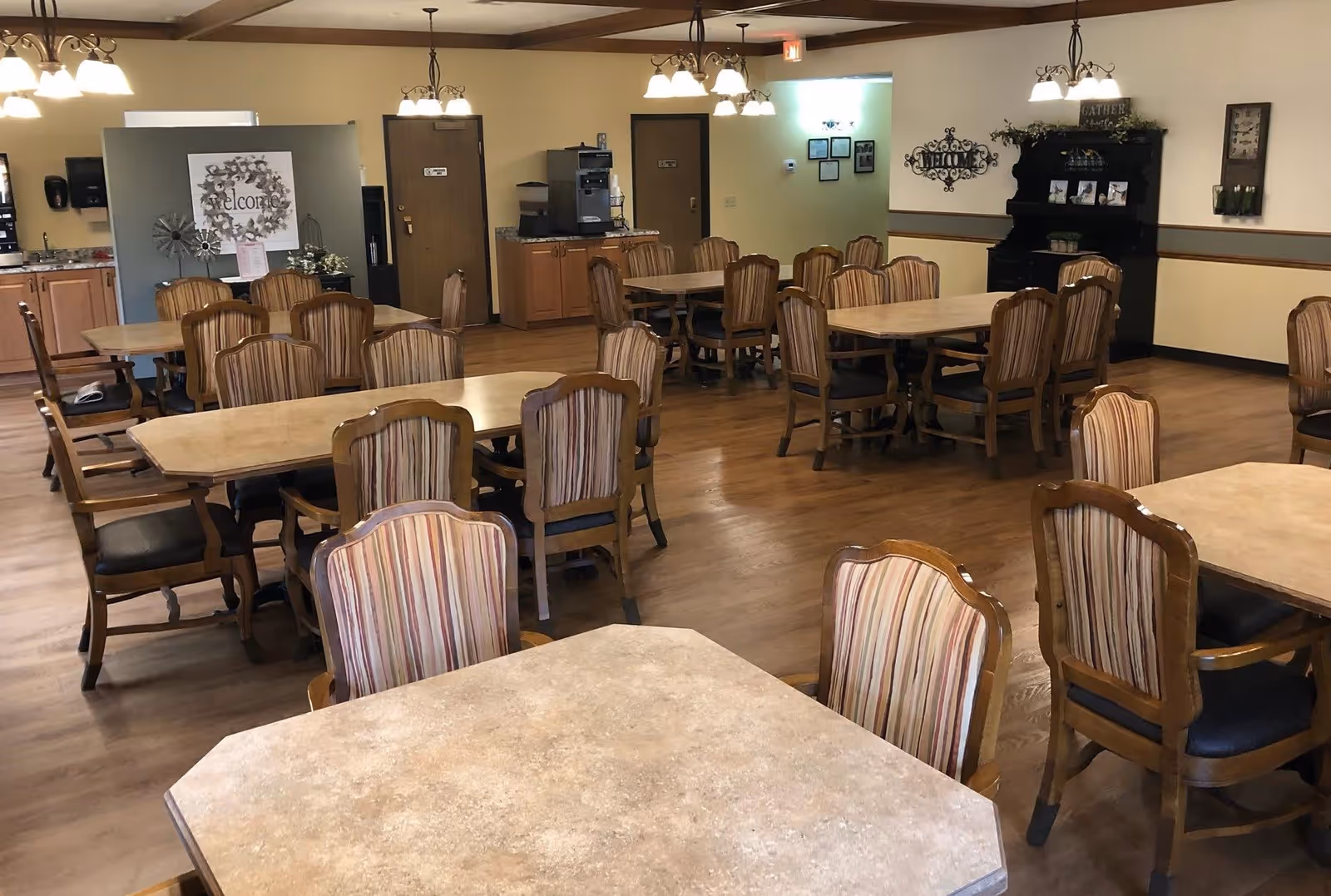 A spacious dining room with multiple wooden tables and chairs arranged neatly. The chairs have striped upholstery. The room features wooden flooring, warm lighting fixtures hanging from the ceiling, and a welcoming decor including a 'Welcome' sign and a black hutch with decorative items. There is a coffee station and some doors in the background.