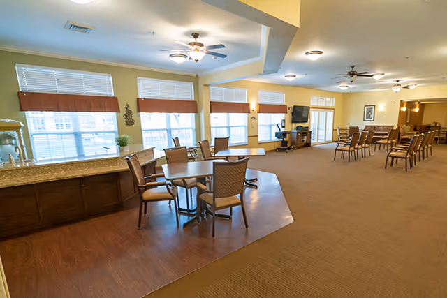 A spacious dining area with multiple tables and chairs arranged neatly. Large windows with brown valances allow natural light to fill the room. There is a counter with a water dispenser on the left side and a television mounted on the wall in the background. Ceiling fans and light fixtures are visible on the ceiling.