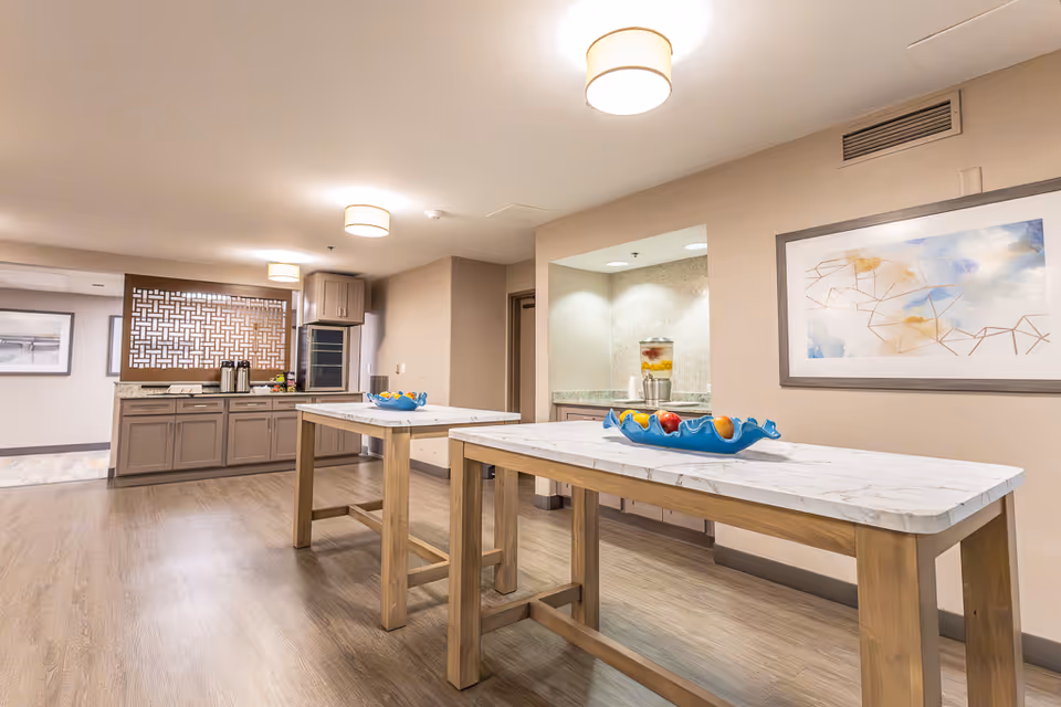 A bright and clean interior space with two wooden tables topped with white marble surfaces, each holding a blue decorative bowl filled with fruit. In the background, there is a counter with cabinets, a coffee station, and a decorative wooden lattice panel. The walls are beige, and there is a framed abstract artwork on the right wall. The floor is light wood, and ceiling lights illuminate the area.