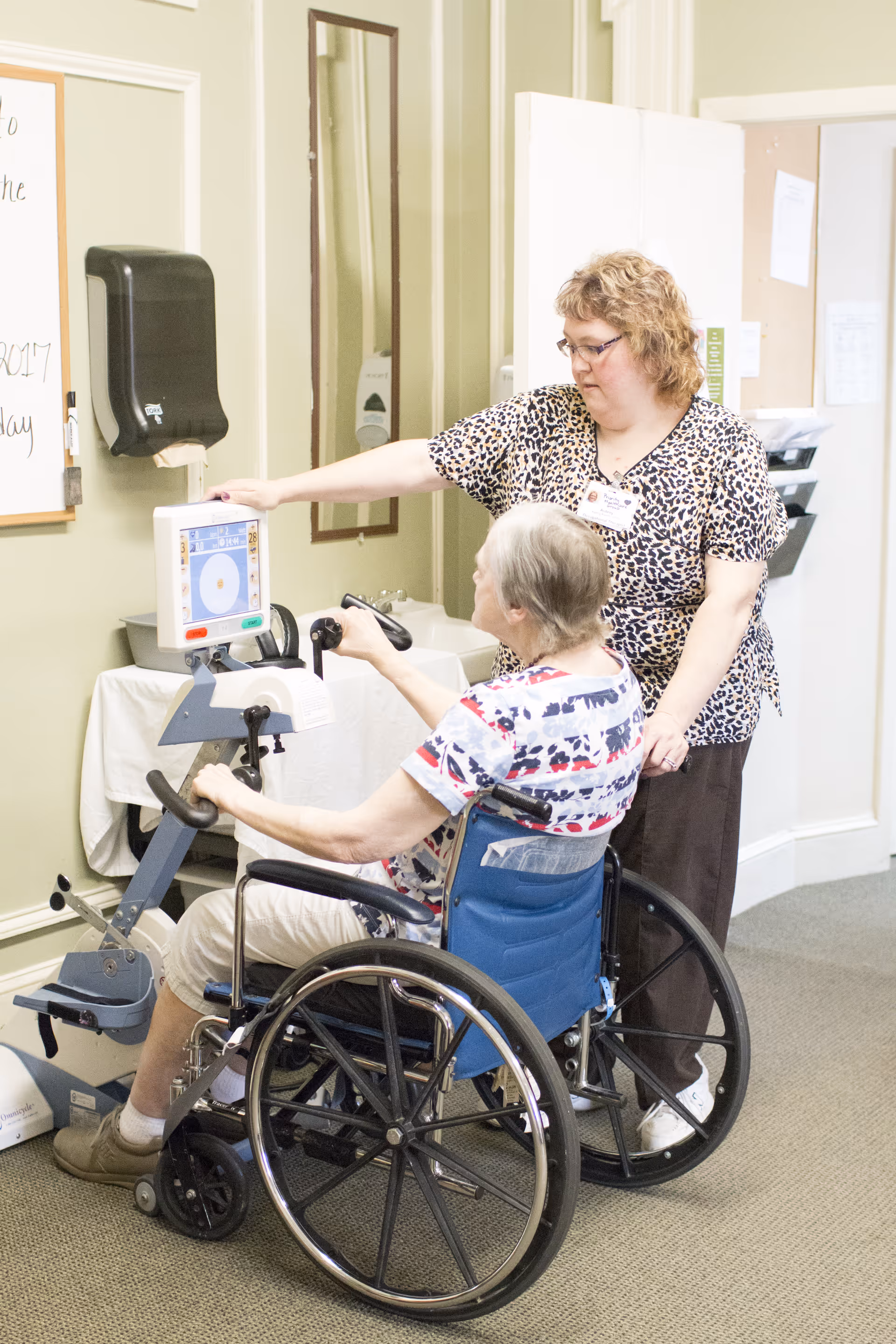 A woman in a wheelchair is using a hand cycle exercise machine while a caregiver stands beside her, assisting and observing. The setting appears to be an indoor rehabilitation or therapy room with light green walls and a bulletin board in the background.