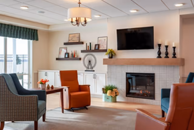 A cozy living room area with a fireplace, a mounted flat-screen TV above it, and decorative candles on the mantel. The room has comfortable armchairs in blue and orange colors arranged around a small wooden table with a flower pot. There are shelves with framed pictures and decorative items on the wall behind the chairs, and large windows with green valances letting in natural light.