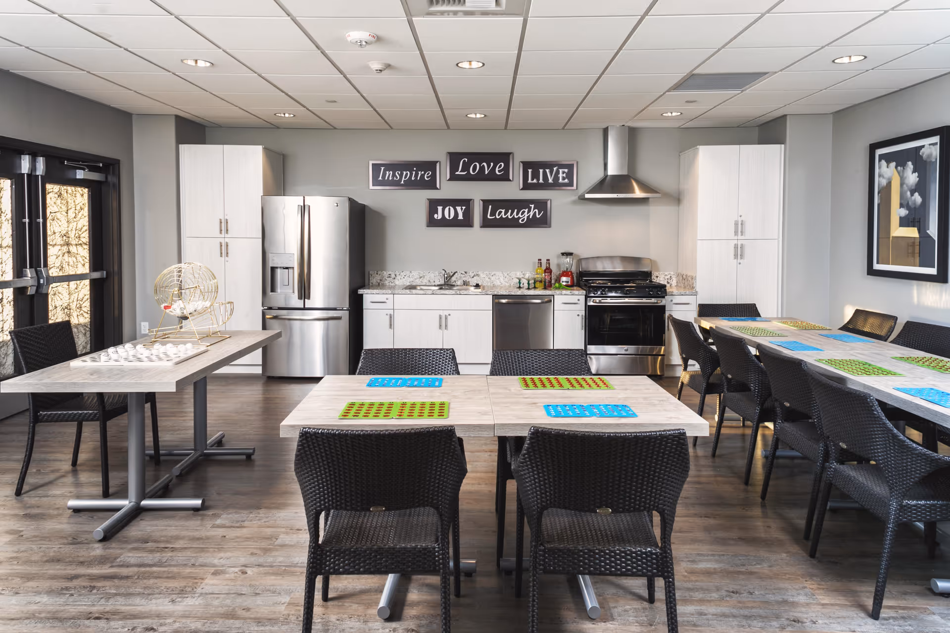 A modern kitchen and dining area with stainless steel appliances including a refrigerator, dishwasher, and stove. There are multiple tables with black woven chairs and colorful placemats. On the wall above the kitchen counter are decorative signs with the words Inspire, Love, Live, Joy, and Laugh. A bingo cage and bingo cards are on one of the tables.