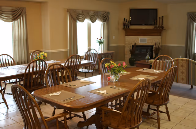 A dining room with wooden tables and chairs arranged neatly. Each table has placemats, napkins, and silverware set for a meal. There are flower vases with colorful flowers on the tables. The room has large windows with beige curtains, and a fireplace with a TV mounted above it is visible in the background.