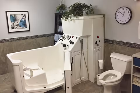 Accessible bathroom featuring a white walk-in assisted bathtub with controls, a toilet, shelving with towels, a clock, and wall art.