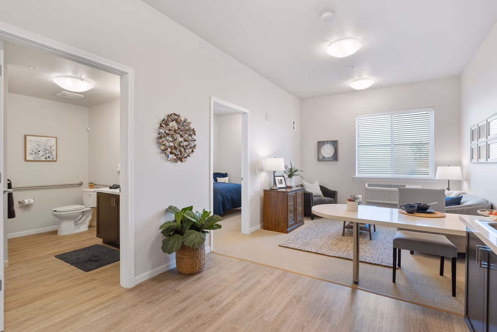 A bright and clean senior living apartment interior featuring a small living room with a sofa, armchair, side table with a lamp, and wall art. Adjacent to the living room is a dining area with a table and chairs. To the left, there is an open doorway leading to a bathroom with a toilet, sink, and framed artwork. Another doorway leads to a bedroom with a blue bedspread. The flooring transitions from wood in the kitchen and hallway to carpet in the living and bedroom areas. A large window with blinds allows natural light into the space.