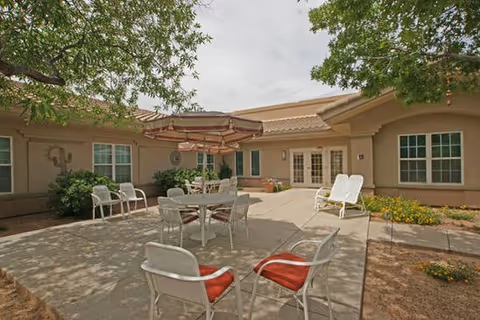 Sunlit courtyard with patio tables, chairs and an umbrella surrounded by a single-story building and trees.