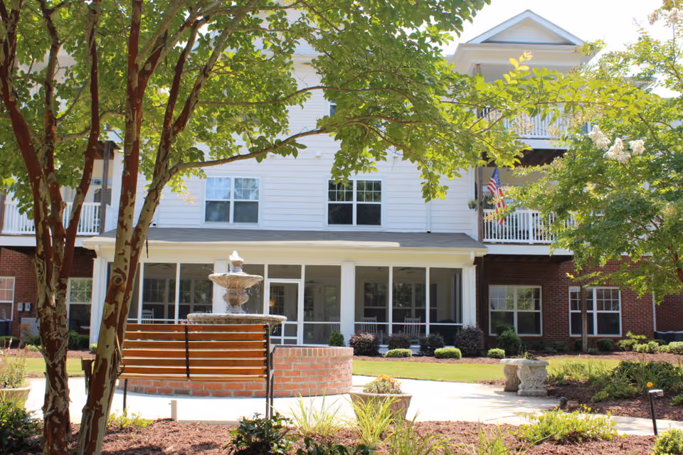 Exterior front of a senior living building showing a fountain, bench, trees, and balconies.