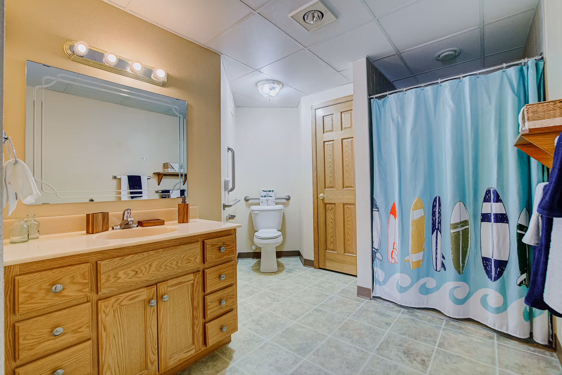 Well-lit accessible bathroom with a wooden vanity and mirror, toilet with grab bar, and a shower with a surfboard-patterned curtain.