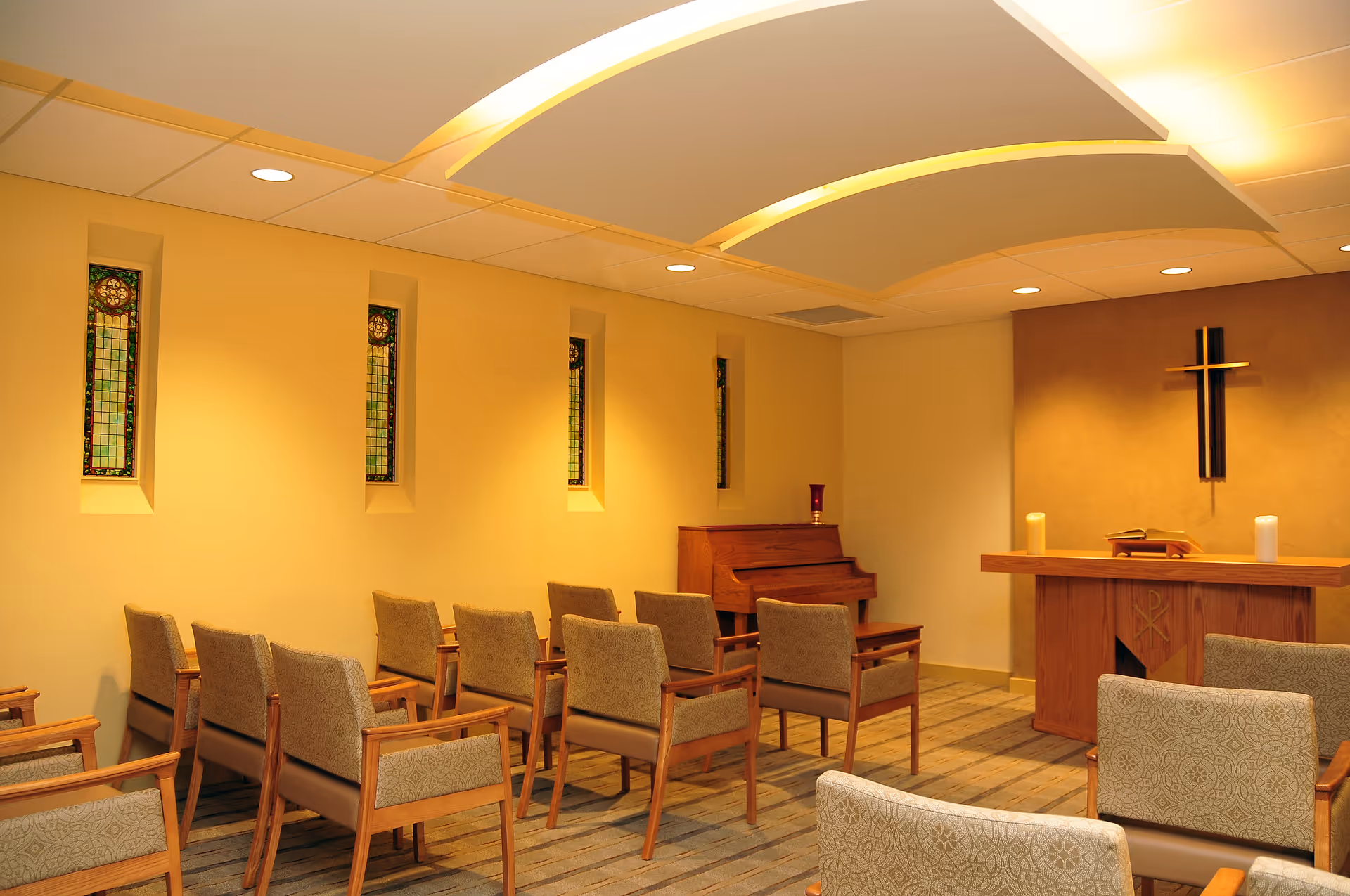 Interior view of a small chapel or meditation room with rows of upholstered chairs facing a wooden altar. The altar has a cross mounted on the wall behind it, two candles, and an open book. There is a piano against the wall with a decorative candle on top. The room has soft lighting and narrow stained glass windows along one wall.