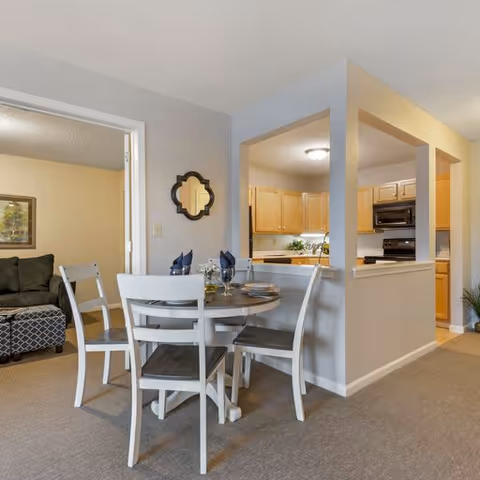 A dining area with a round wooden table set for four with plates, glasses, and napkins. The dining area is adjacent to a kitchen with wooden cabinets and black appliances. In the background, there is a living room with a dark green sofa and a patterned ottoman. The walls are painted light gray and beige, and the floor is carpeted.