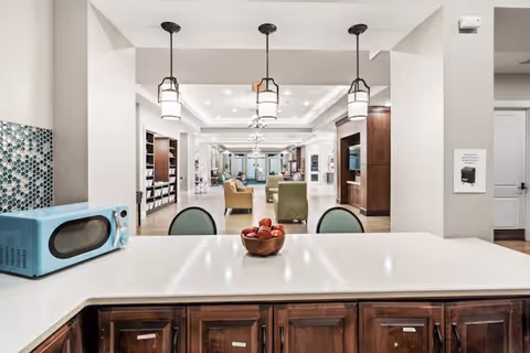 View from a kitchen counter with a bowl of red apples and a blue microwave on the left side, looking out into a spacious, well-lit living area with chairs, bookshelves, and pendant lights hanging from the ceiling.