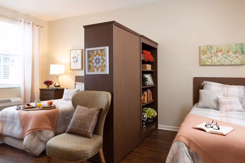 A cozy bedroom in a senior living facility with two beds separated by a tall wooden bookshelf. Each bed has light-colored bedding with peach blankets. A cushioned chair with a decorative pillow is in the foreground. The room is softly lit by a table lamp on a nightstand next to one bed, and there are framed artworks on the walls. A tray with breakfast items is placed on one bed, and a pair of reading glasses rests on an open book on the other bed.