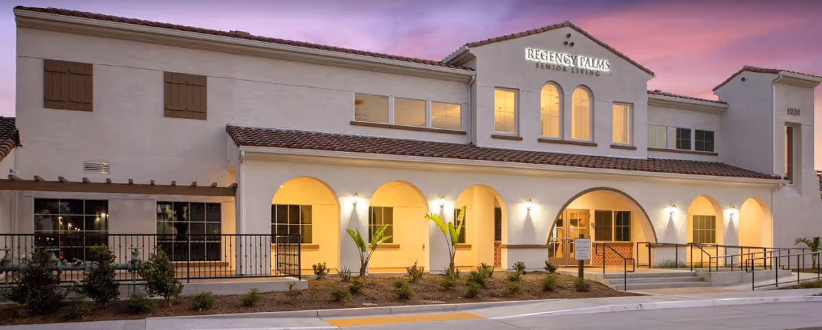 Exterior front facade of a two-story senior living building with arched entryways and illuminated windows at dusk.