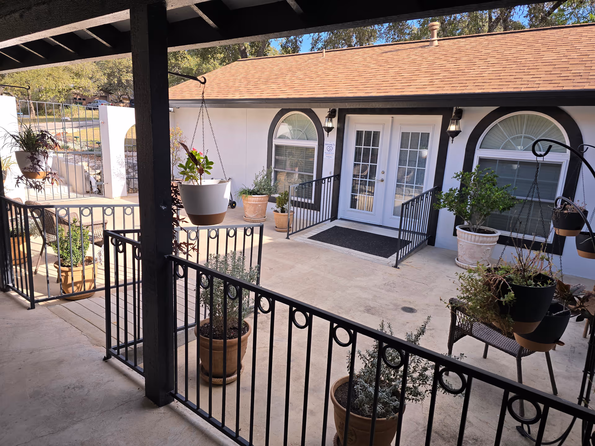 Outdoor patio area with potted plants and hanging planters, black metal railings, and a white building with arched windows and double glass doors in the background.