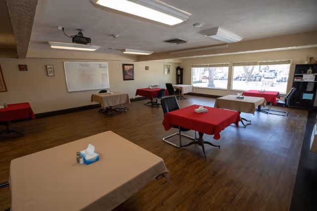 A spacious room with multiple tables covered in red and beige tablecloths, each with a tissue box and salt and pepper shakers. The room has large windows letting in natural light, a whiteboard on one wall, and a wooden floor. Several chairs are placed around the tables, and there is a bookshelf and a grandfather clock in the background.