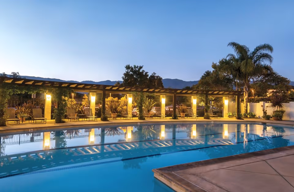 Outdoor swimming pool at dusk with a pergola along one side featuring illuminated columns and several lounge chairs. Palm trees and other plants surround the area, with mountains visible in the background under a clear sky.