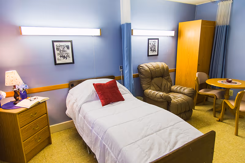 A cozy bedroom in a senior living facility featuring a single bed with white bedding and a red pillow, a wooden nightstand with a lamp and flowers, a comfortable recliner chair, a wooden wardrobe, and a small round table with three chairs. The walls are painted light blue with framed artwork and a fluorescent light fixture above the bed.
