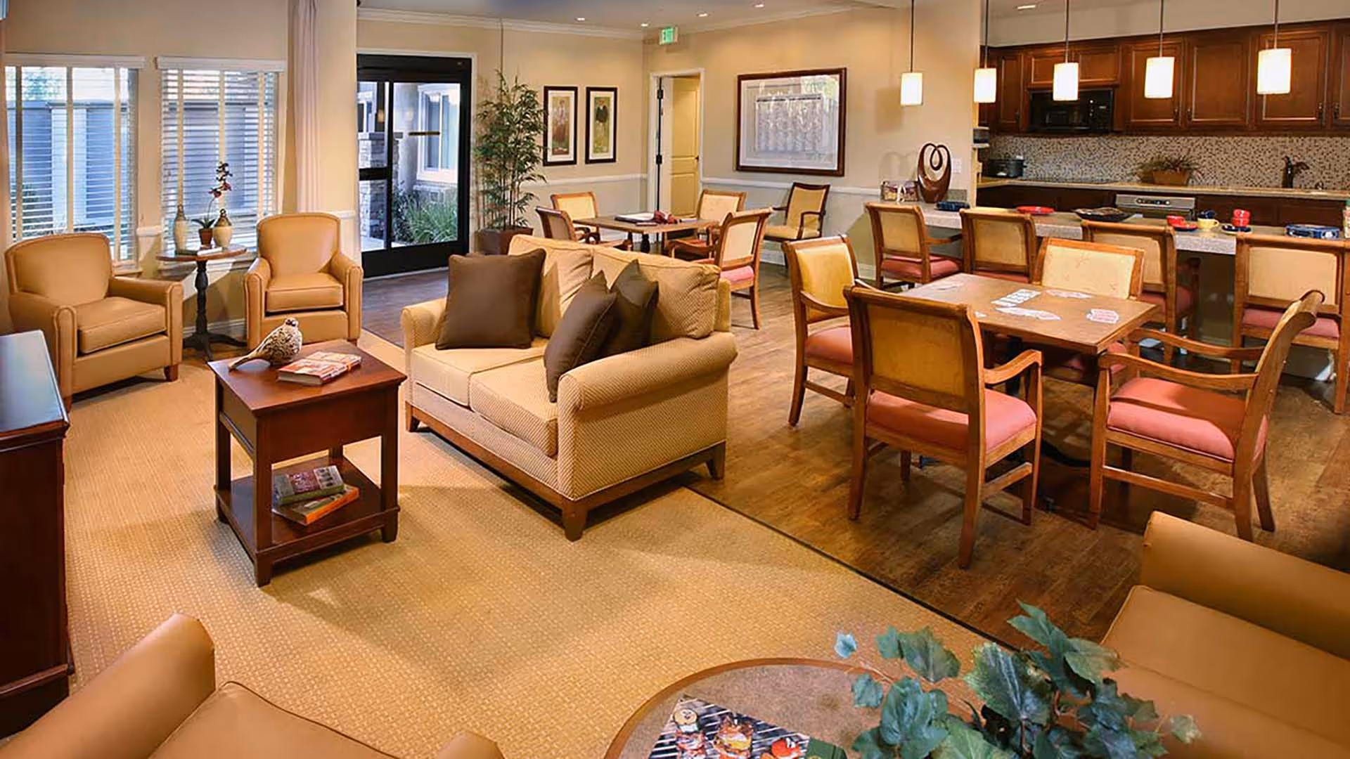 A cozy and well-lit common area in a senior living facility featuring a beige sofa with brown pillows, several armchairs, a wooden coffee table with books and decorative items, multiple dining tables with chairs, and a kitchen area with hanging pendant lights and wooden cabinets in the background.