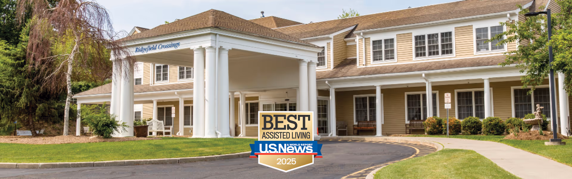 Exterior view of Benchmark at Ridgefield Crossings, showing a large building with beige siding, white columns, and a covered entrance. There is a curved driveway and a sidewalk leading to the entrance, with green grass and some trees around the building. A sign in front reads 'Best Assisted Living U.S. News 2025'.