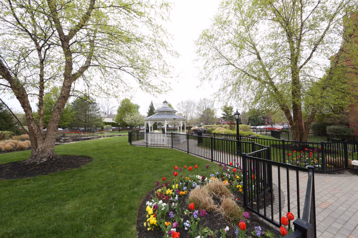 A well-maintained outdoor garden area with green grass, colorful flower beds including tulips and pansies, and several trees with fresh green leaves. A paved walkway with black metal railings leads to a white gazebo in the center. There are lamp posts along the path and a parking lot with cars visible in the background.