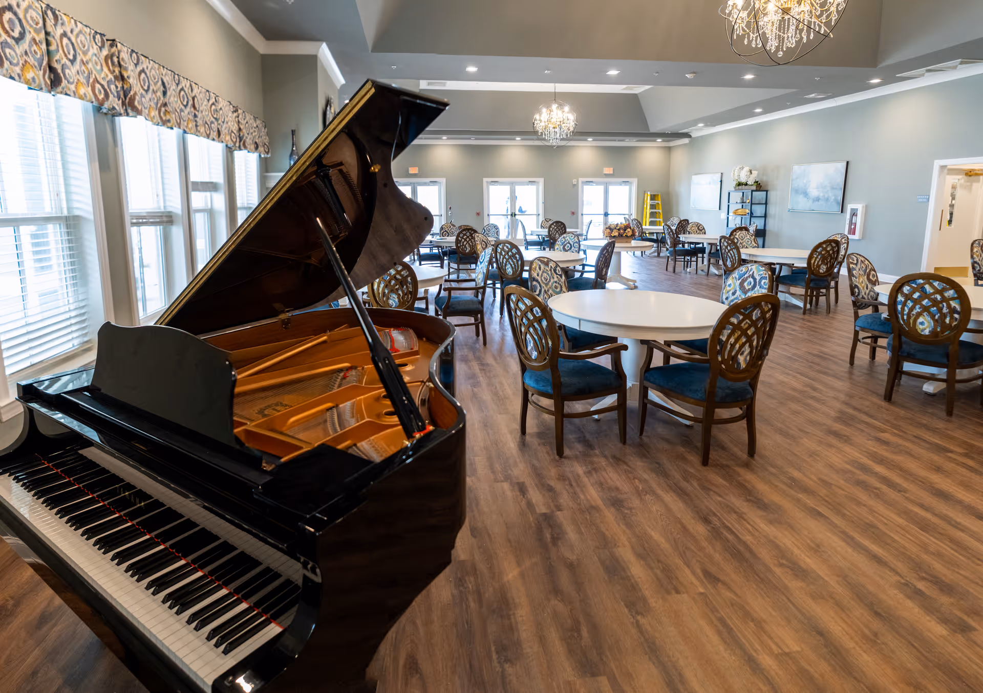Spacious dining room with multiple round tables and patterned chairs on a wooden floor. A grand piano with its lid open is positioned near large windows with patterned valances. The room is well-lit with chandeliers and has light-colored walls with artwork and shelves in the background.