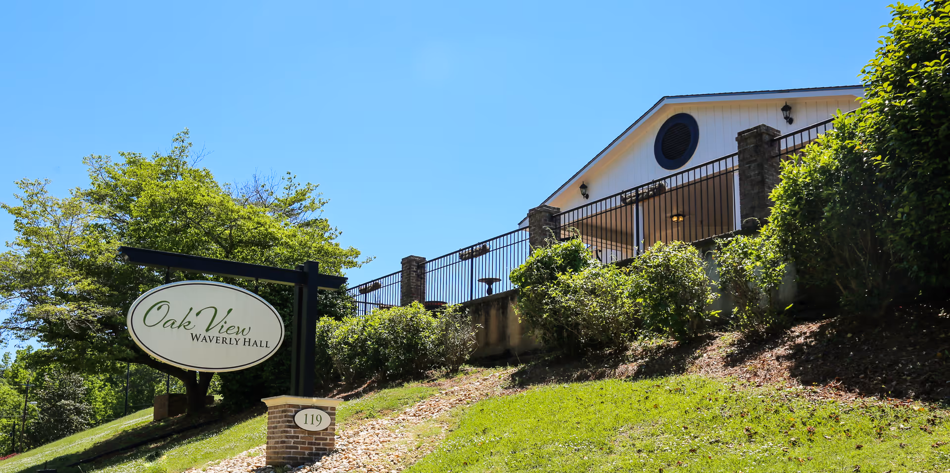 Exterior view of Oak View Waverly Hall facility on a sunny day, showing a green lawn, bushes, a sign with the facility name, and part of the building with a porch and railing.